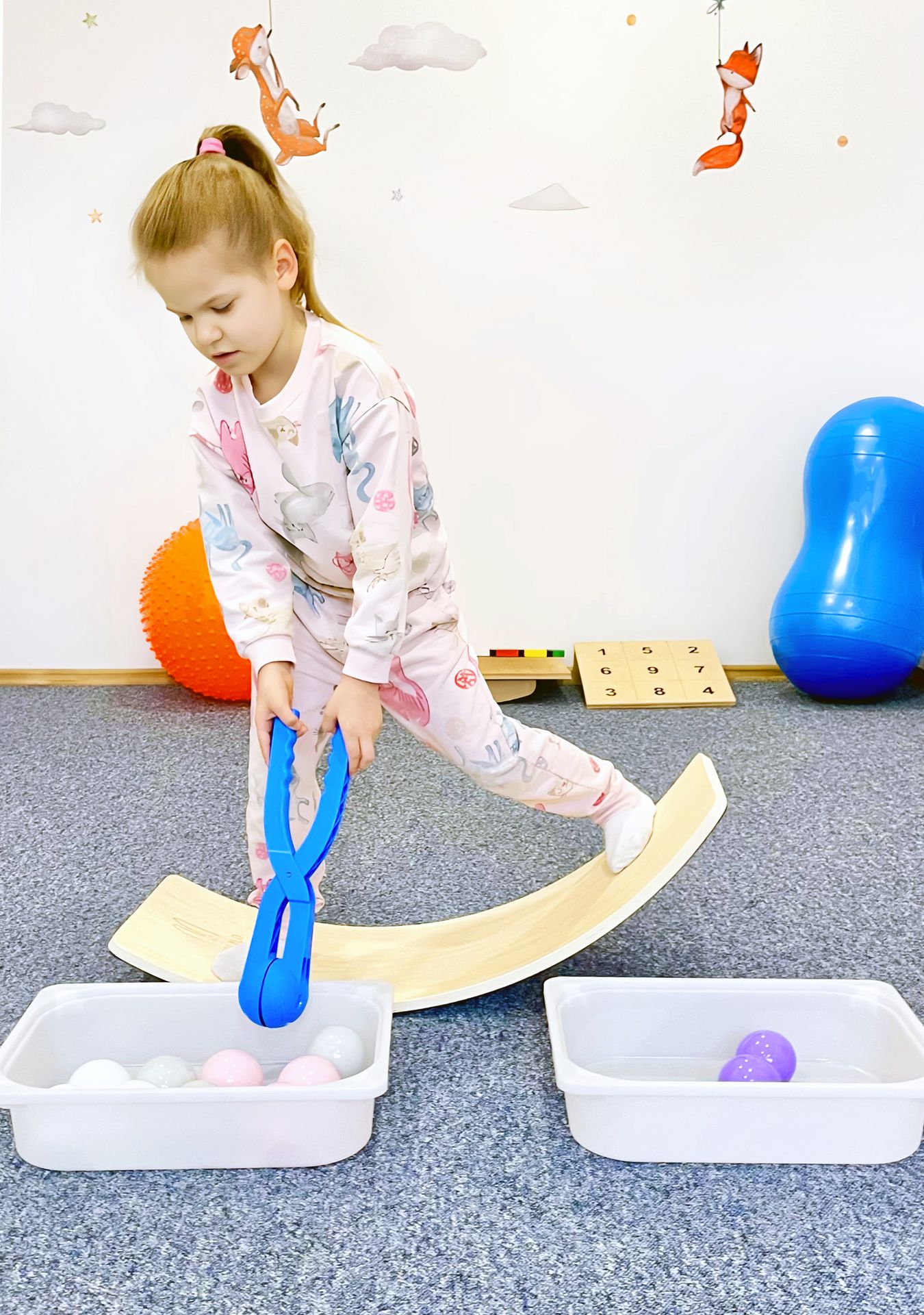 Złożone ćwiczenie koordynacyjne – przenoszenie przedmiotów w niestabilnej pozycji rozwija samoregulację i precyzję ruchów Dziecko przenosi piłki szczypcami, stojąc na balansie, podczas terapii sensorycznej w Centrum Wzlot