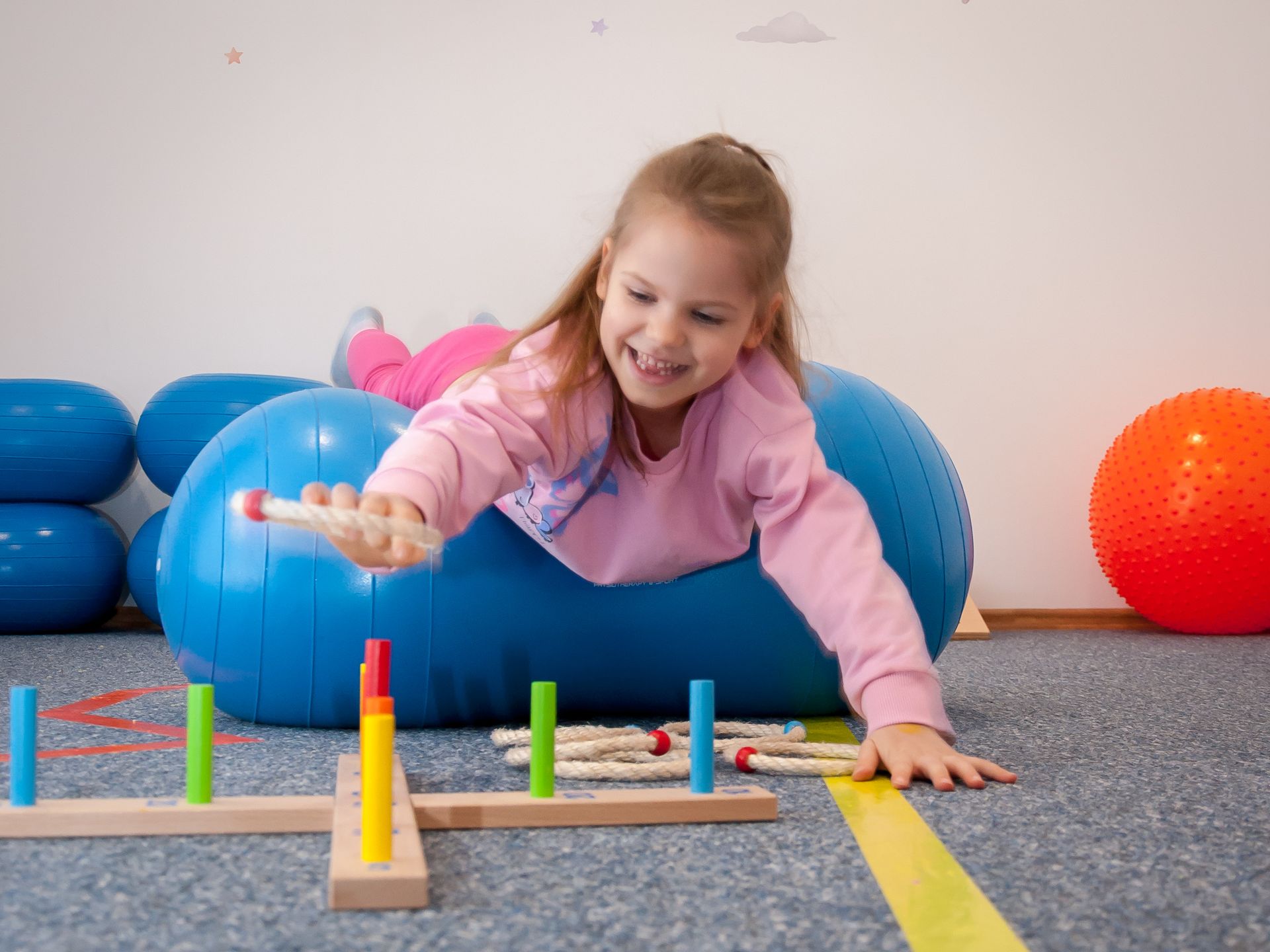 Radosne i rozwijające ćwiczenie: dziecko przekłada elementy leżąc na piłce fasolce. Wzmacnia to koordynację ruchową, siłę i małą motorykę. Dziecko wykonuje ćwiczenie na piłce fasolce w Centrum Rozwoju Wzlot w Krakowie