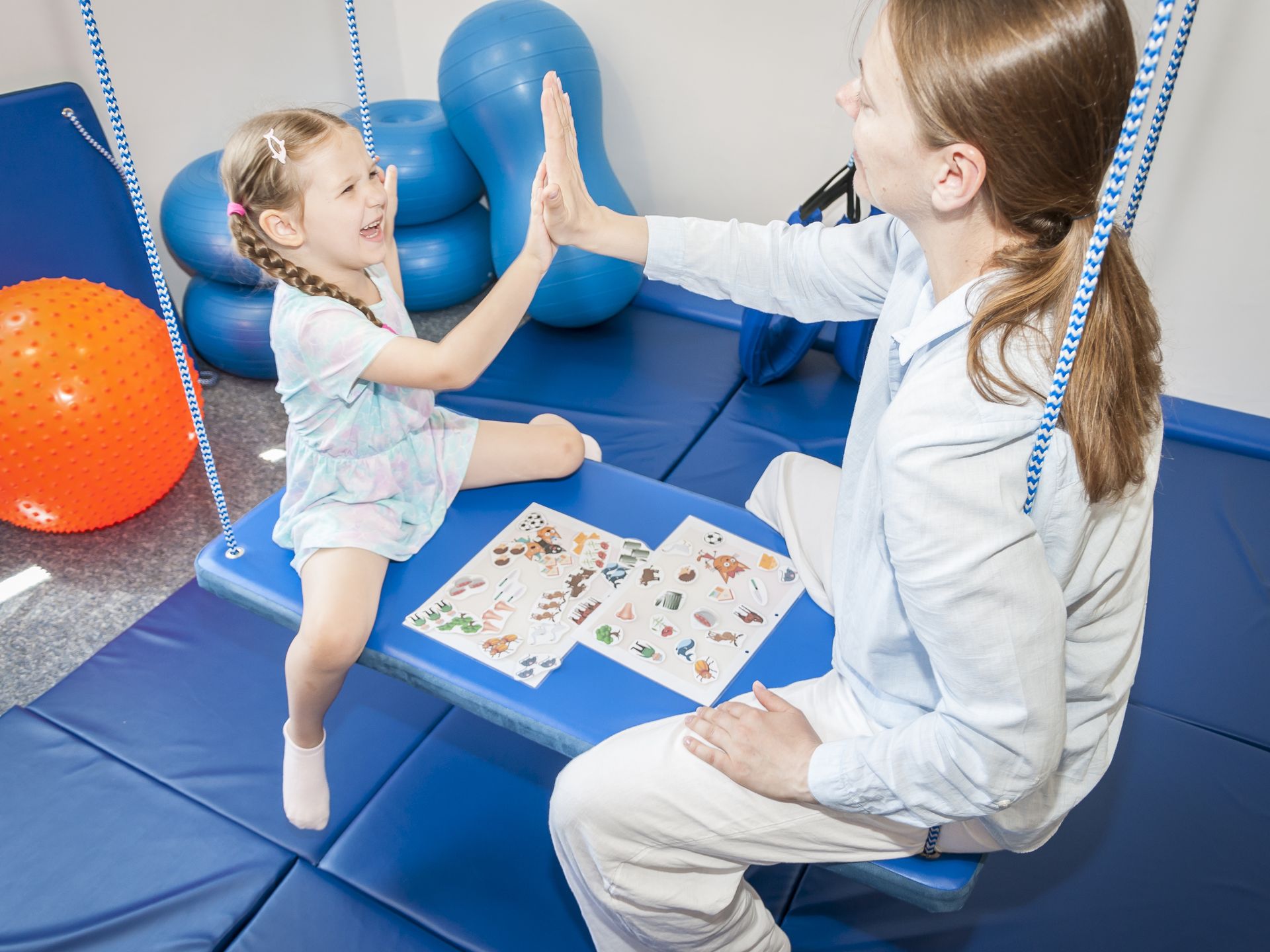 Движение и мотивация помогают речи Девочка на подвесных качелях даёт пять терапевту на занятии